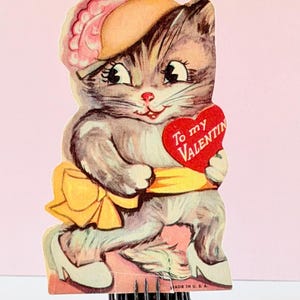 May include: Vintage Valentine's Day card featuring a gray and white cat holding a red heart with the text "To my Valentine." The cat wears a pink hat and yellow bow. The card is mounted on a metal floral frog.