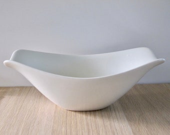 May include: A white, oblong ceramic bowl with a curved, boat-like shape. The bowl has a smooth, matte finish and is set on a light-colored wooden surface. The design features gently flared sides and a wide opening.