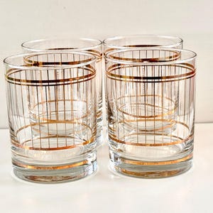 May include: Four clear drinking glasses with a gold grid pattern. The glasses have a cylindrical shape with horizontal gold bands at the top, middle, and bottom. The gold grid design adds a vintage touch to the glassware.