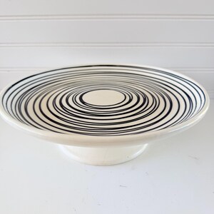 May include: A white ceramic cake stand. The plate features a black spiral design. The stand has a circular base and a raised edge. The black lines create a concentric pattern. The stand is approximately 20 cm in diameter.