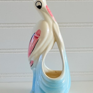 May include: A ceramic flamingo figurine in cream, pink, and light blue. The bird has a long neck and a curved body, with a small opening in the center. The flamingo's beak and wing details are painted in pink and black.