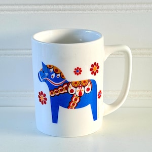 May include: White ceramic mug featuring a blue horse design with decorative patterns and red flower accents. The mug has a curved handle and a cylindrical shape, suitable for hot beverages.