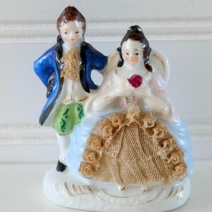 May include: A porcelain figurine of a couple standing together. The figure on the left wears a blue coat, green breeches, and a tan cravat. The figure on the right wears a light blue dress with a tan lace overlay and holds a small red flower.