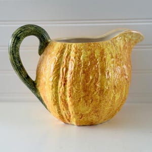 May include: A ceramic pitcher shaped like a pumpkin with a textured yellow and orange body and a dark green handle. The vessel features a rounded, ribbed exterior with a wide spout and a curved handle that mimics the appearance of a pumpkin stem.