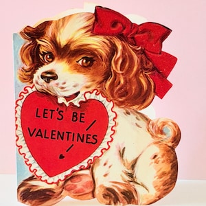 May include: Vintage Valentine's Day card featuring a Cavalier King Charles Spaniel dog with brown and white fur, wearing a red bow. The dog holds a red heart with the text "LET'S BE VALENTINES" on a pink background.