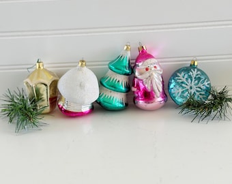 Peut inclure: Un ensemble de cinq décorations de Noël en verre vintage comprenant une lanterne dorée, un champignon blanc, un sapin vert, une figurine de Père Noël rose et une sphère bleue avec un flocon de neige à paillettes blanches, avec des brins de pin.