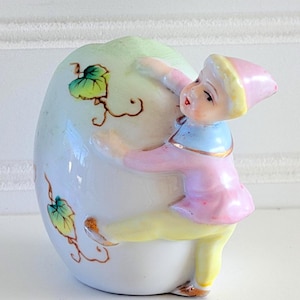 May include: A vintage ceramic figurine of a small figure in pink and yellow clothing, hugging a large, egg-shaped object. The egg is white with green and brown leaf accents. The figure has a pink hat and blue collar.