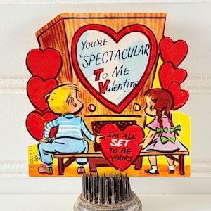 May include: A vintage Valentine's Day card with a retro TV design and a heart-shaped screen. The card reads "You're Spectacular To Me Valentine" and "I'm All Set To Be Yours". It is mounted on a metal floral frog, a common florist's tool.