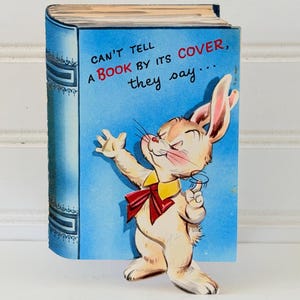 以下が含まれることがあります： 赤い蝶ネクタイと黄色の襟を着た漫画のウサギが描かれた、本型のヴィンテージグリーティングカード。青い表紙には「CAN'T TELL A BOOK BY ITS COVER, they say...」と書かれています。