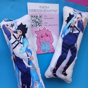 Puede incluir: Dos llaveros de peluche rectangulares con ilustraciones de personajes estilo anime en trajes azules, blancos y negros. Entre ellos hay una tarjeta blanca con el texto FATH OBESSIVEARTIST, un código QR y una pequeña ilustración de dibujos.