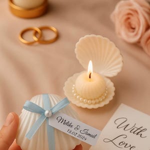 Può includere: Un portacandele a forma di conchiglia con una candela accesa, una base bordata di perle e una piccola candela. Una seconda candela a forma di conchiglia è tenuta con un nastro blu e un'etichetta che recita "Melike & Ismail 13.02.2024". Una carta recita "With Love".