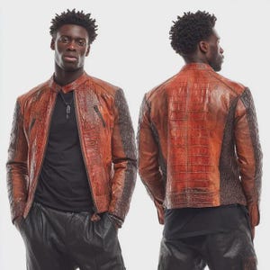 May include: A brown leather jacket with a textured crocodile pattern. The jacket has a zip-up front, a stand-up collar, and dark brown panels on the shoulders and sleeves. The model is wearing a black shirt and black pants.