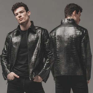 May include: A black, textured leather jacket with a crocodile pattern. The jacket has a zipper closure and a collared neckline. The model is wearing a black shirt and black pants. The jacket is a stylish outerwear piece.