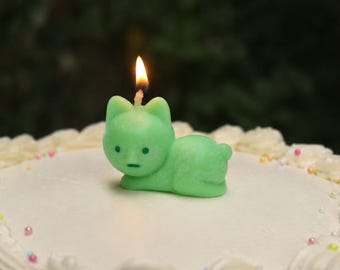 Könnte beinhalten: Eine hellgrüne, katzenförmige Kerze mit brennendem Docht, die auf einem weiß glasierten Kuchen steht. Die Kerze hat einen einfachen Gesichtsausdruck mit zwei kleinen Punkten als Augen und einem horizontalen Strich als Mund.