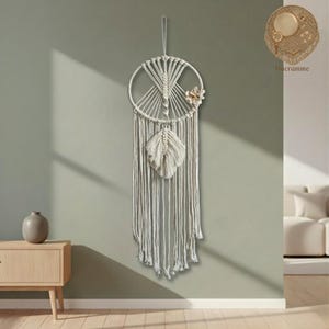 Op de afbeelding: Een crèmekleurige macramé wandhanger met een cirkelvormig ontwerp, met een centrale veer en decoratieve accenten. Het stuk is opgehangen aan een lus en hangt tegen een neutrale muur, wat een vleugje bohemien decor toevoegt.