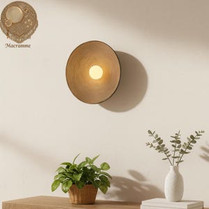 Puede incluir: Aplique de pared redondo, tejido, de color marrón claro, con una fuente de luz central. La superficie es texturizada. La luz es de color cálido. La palabra "Macramme" está impresa en la pared.