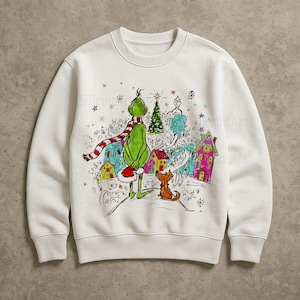 Puede incluir: Sudadera blanca con una colorida ilustración de un personaje verde con una bufanda a rayas rojas y blancas, un gorro rojo y un perro marrón pequeño. El gráfico incluye un árbol de Navidad y casas caprichosas.