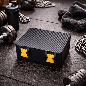 Puede incluir: Una caja de almacenamiento negra con un patrón de fibra de carbono en la tapa y cierres amarillos brillantes. La caja está rodeada de equipos de fitness, incluyendo pesas, una botella mezcladora y una cadena.