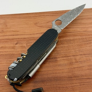 May include: A multi-tool pocket knife featuring a patterned Damascus steel blade with a circular cutout, black textured handle scales, a corkscrew, and a metallic pen attachment. A black lanyard with a small rectangular end piece is attached to the base.