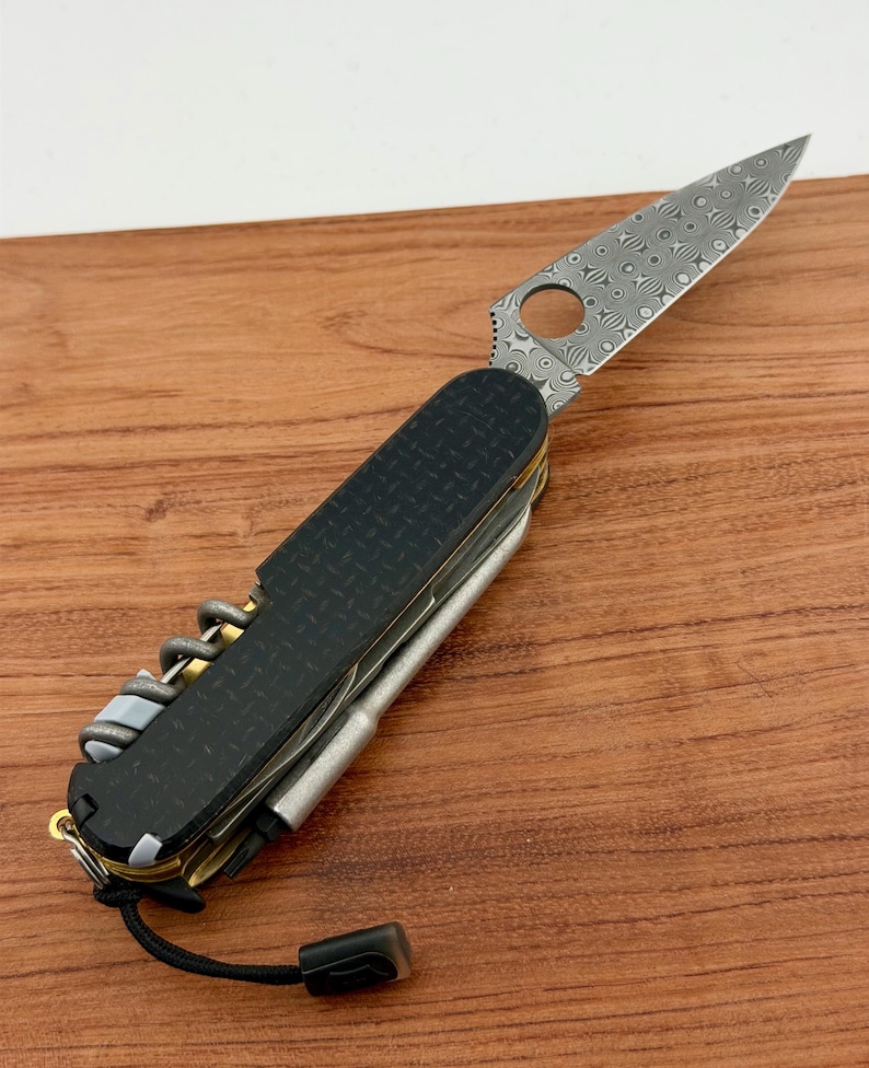 May include: A multi-tool pocket knife featuring a patterned Damascus steel blade with a circular cutout, black textured handle scales, a corkscrew, and a metallic pen attachment. A black lanyard with a small rectangular end piece is attached to the base.