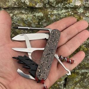 May include: A multi-tool pocket knife with a patterned copper and black handle. The knife features various tools, including a screwdriver, scissors, and a corkscrew. A red lanyard is attached to the base of the knife.