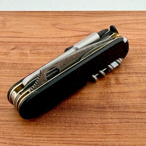 May include: A multi-tool pocket knife featuring black textured handle scales, brass liners, and a Damascus steel blade. The tool includes a silver-toned screwdriver bit, a corkscrew, and various integrated metal implements folded into the frame.