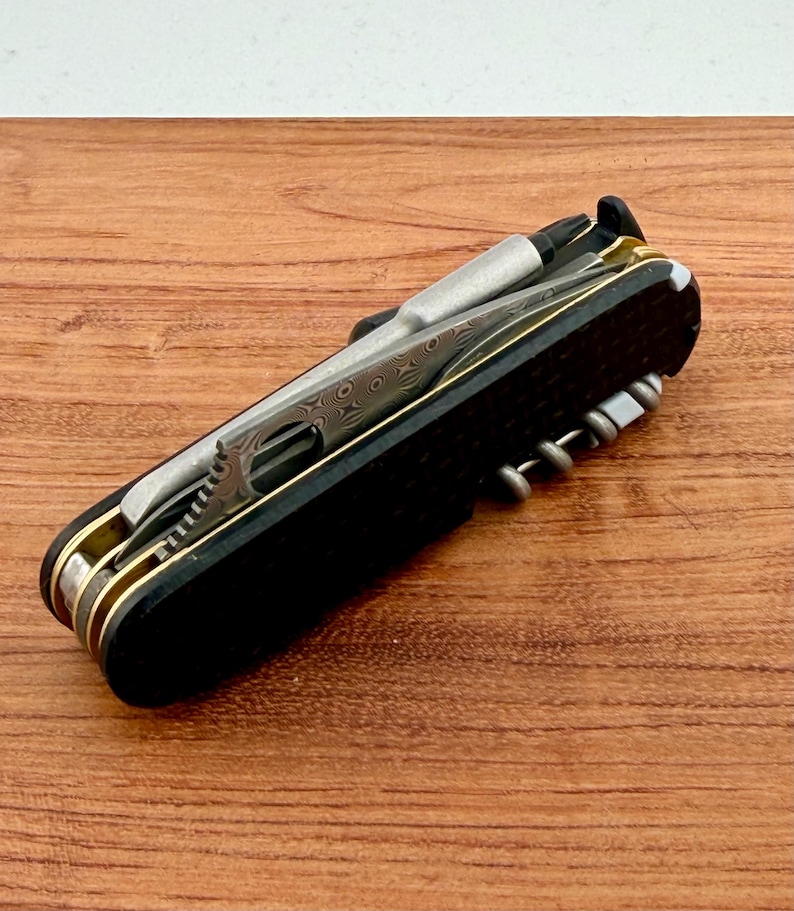 May include: A multi-tool pocket knife featuring black textured handle scales, brass liners, and a Damascus steel blade. The tool includes a silver-toned screwdriver bit, a corkscrew, and various integrated metal implements folded into the frame.