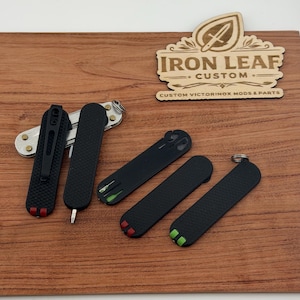 May include: Several black custom Victorinox mods and parts are displayed on a wooden surface. The parts include pocket clips and scales with red and green accents. The wooden sign reads "Iron Leaf Custom" and "Custom Victorinox Mods & Parts."