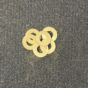 Puede incluir: Un grupo de seis arandelas pequeñas de color dorado. Cada arandela tiene forma circular con un agujero en el centro. Las arandelas están dispuestas en un patrón ligeramente superpuesto sobre un fondo gris oscuro texturizado.