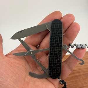 May include: A multi-tool with a black textured handle and various tools extended, including a knife blade, scissors, corkscrew, and bottle opener. The tools are made of a silver-colored metal. A black lanyard is attached to the base.