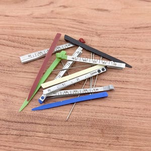Könnte beinhalten: Eine Sammlung farbenfroher Nähwerkzeuge, darunter Nadeln und Messwerkzeuge. Die Werkzeuge sind in verschiedenen Farben gehalten, darunter Grün, Rot, Schwarz und Blau. Die Messwerkzeuge sind mit Zollmaßen versehen.