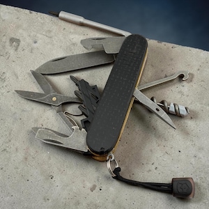 May include: A multi-tool pocket knife with a black textured handle featuring a Swiss cross emblem. The tool is fully extended, revealing various stainless steel implements including a blade, scissors, screwdriver, corkscrew, and a hook.