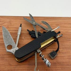 May include: A multi-tool pocket knife featuring a Damascus steel blade with a circular cutout, a black textured handle, and various deployed implements including scissors, a corkscrew, a metal pick, and a small black lanyard with a logo tag.