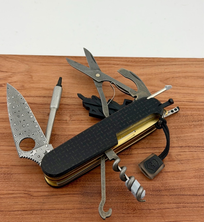 May include: A multi-tool pocket knife featuring a Damascus steel blade with a circular cutout, a black textured handle, and various deployed implements including scissors, a corkscrew, a metal pick, and a small black lanyard with a logo tag.