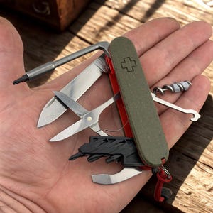 May include: A multi-tool with a green and red handle, featuring various tools like a knife, scissors, and screwdriver. The tool has a cross emblem and a red lanyard. The tools are open, displaying their functionality.