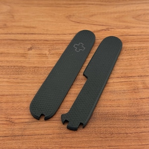 May include: Two black replacement scales for a pocket knife, featuring a textured surface and rounded ends. One scale displays a small embossed Swiss cross emblem near the top, while the other includes a specific cutout notch along the side edge.