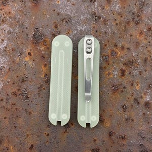 May include: Two pale green, rectangular knife handle scales with rounded ends. One scale features a silver metal pocket clip attached with two screws. Both scales have a textured surface and two circular recessed holes at each end.
