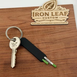May include: A black and silver key with a black handle and a small ruler with green accents. The key is attached to a silver keyring. The wooden sign reads "Iron Leaf Custom" and "Custom Victorinox Mods & Parts."