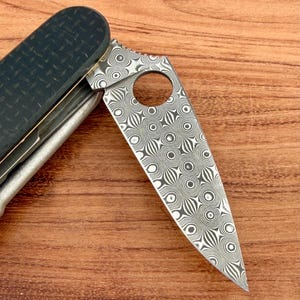 May include: A folding pocket knife featuring a blade with a distinct circular Damascus steel pattern and a circular cutout near the spine. The handle is constructed from a dark, textured carbon fiber material with a visible weave pattern.
