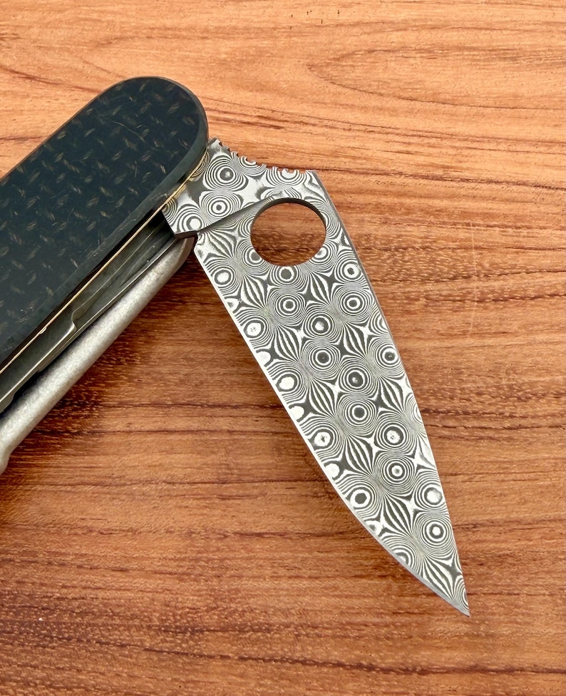 May include: A folding pocket knife featuring a blade with a distinct circular Damascus steel pattern and a circular cutout near the spine. The handle is constructed from a dark, textured carbon fiber material with a visible weave pattern.