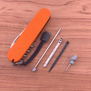 Könnte beinhalten: Ein orangefarbenes Schweizer Taschenmesser mit einem Kreuzemblem, zusammen mit mehreren Werkzeugen, darunter ein Metalllineal, ein Zahnstocher und ein kleiner Schraubendreher. Die Werkzeuge liegen auf einer Holzoberfläche.