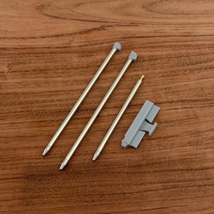 Puede incluir: Tres lápices ópticos plateados con puntas grises y un clip de plástico gris se muestran sobre una superficie de madera. Los lápices tienen un diseño metálico elegante y el clip tiene un área pequeña y texturizada.