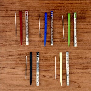 May include: An overhead shot shows a collection of small, colorful tools and measuring devices. There are red, blue, green, black, and cream-colored tools, each paired with a silver ruler marked in millimeters. The tools are arranged on a wooden surface.