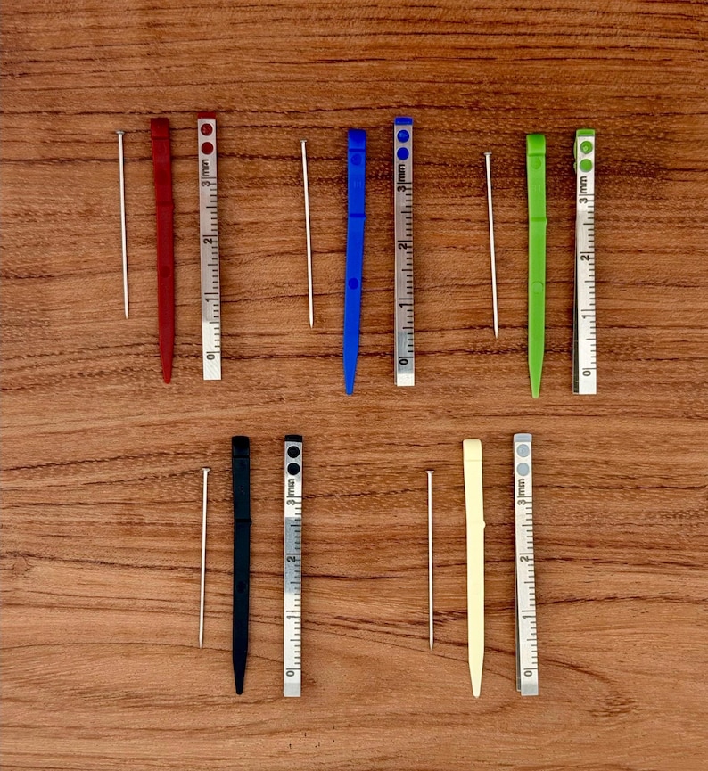 May include: An overhead shot shows a collection of small, colorful tools and measuring devices. There are red, blue, green, black, and cream-colored tools, each paired with a silver ruler marked in millimeters. The tools are arranged on a wooden surface.