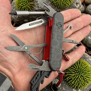 May include: A multi-tool pocket knife featuring a black textured handle with a Swiss cross emblem and a bright red interior frame. The tool includes multiple extended stainless steel implements such as scissors, a blade, a screwdriver, and a hook.