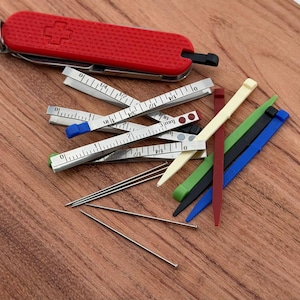Op de afbeelding: Een rood Zwitsers zakmes met een kruisembleem, zilveren vouwlinialen en kleurrijke plastic gereedschappen zijn gerangschikt op een houten oppervlak. Er zijn ook verschillende zilveren spelden. De gereedschappen zijn voor knutselen of naaien.