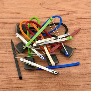 Op de afbeelding: Een verzameling kleurrijke, miniatuur meetgereedschappen met bevestigde koorden. De gereedschappen omvatten linialen gemarkeerd in inches en millimeters, met diverse gekleurde accenten zoals groen, blauw, oranje en rood. De gereedschappen zijn gerangschikt op een houten oppervlak.