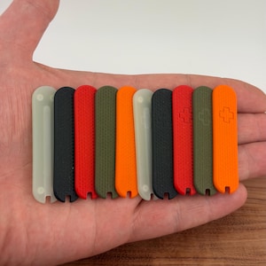 May include: A collection of ten multi-colored replacement scales for a pocket knife. The scales are in various colors, including black, red, orange, and green. Each scale has a textured surface and a small notch at the bottom.
