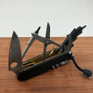 May include: A multi-tool pocket knife with a black textured handle and gold-colored interior. It features a Damascus steel blade with a circular cutout, a metal bit driver, scissors, a bottle opener, and a black lanyard with a small logo tag.