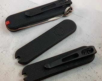 May include: Three black pocket knife scales with a textured surface. Two scales feature a black metal pocket clip attached with screws, while one scale displays a recessed cross emblem. One scale is attached to a folded knife with a red accent.